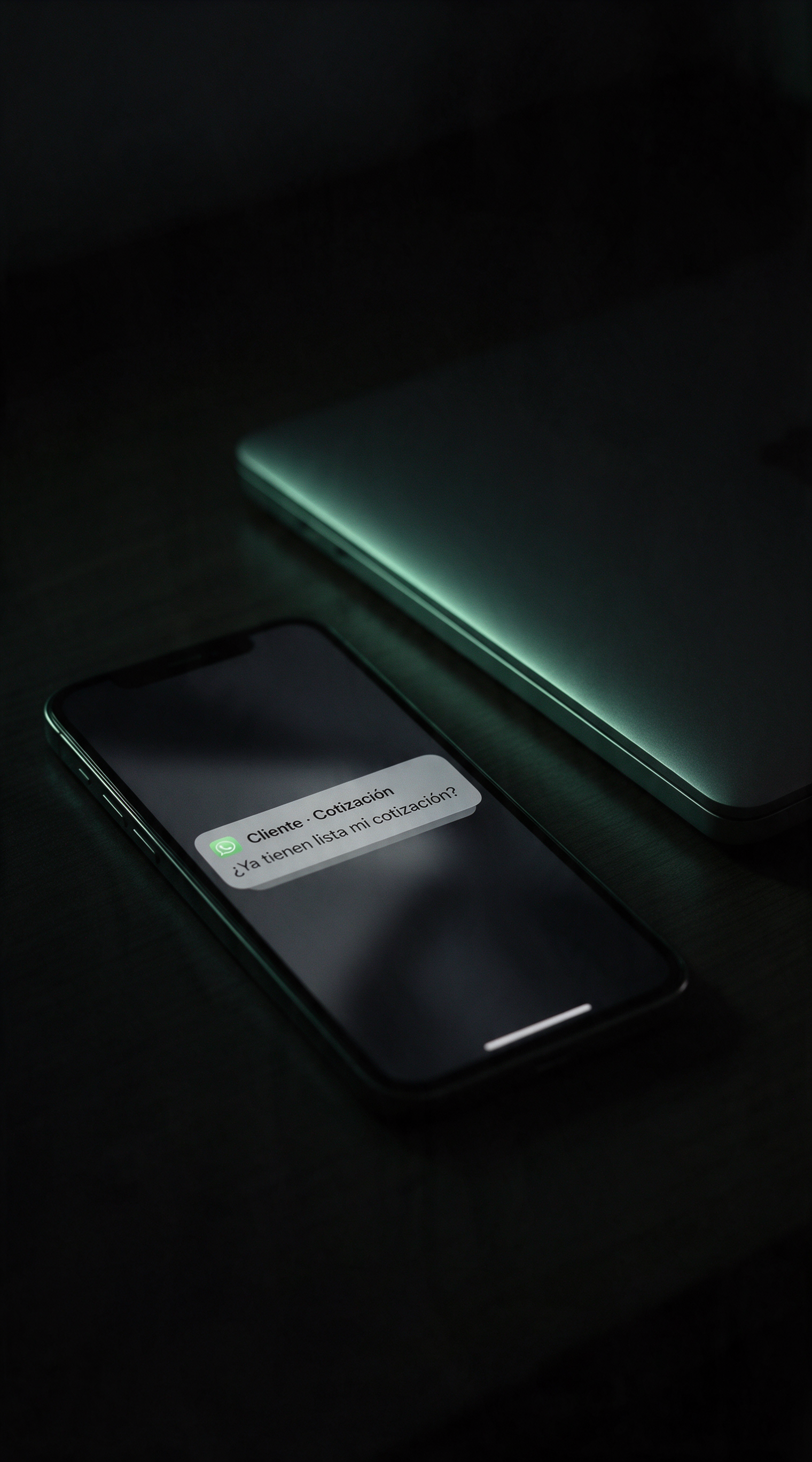 Un teléfono y una laptop cerrada sobre un escritorio de madera, en un cuarto oscuro de noche.